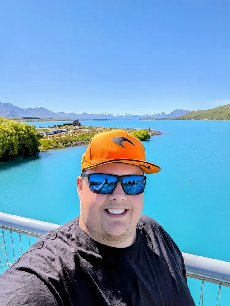 Ben smiling in an orange cap and sunglasses, with turquoise Lake Tekapo and snow-capped mountains behind him.